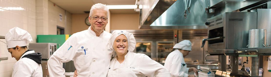 A Culinary Arts student and chef stand smiling in a Pima Culinary Kitchen