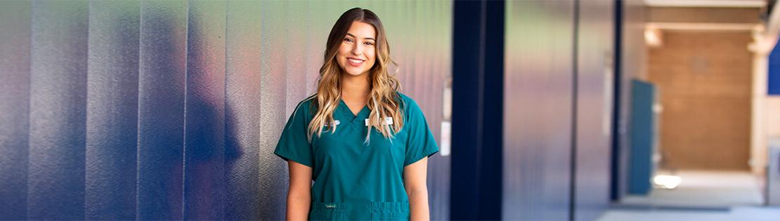 A Pima nursing student stands smiling against a wall at Pima's West Campus
